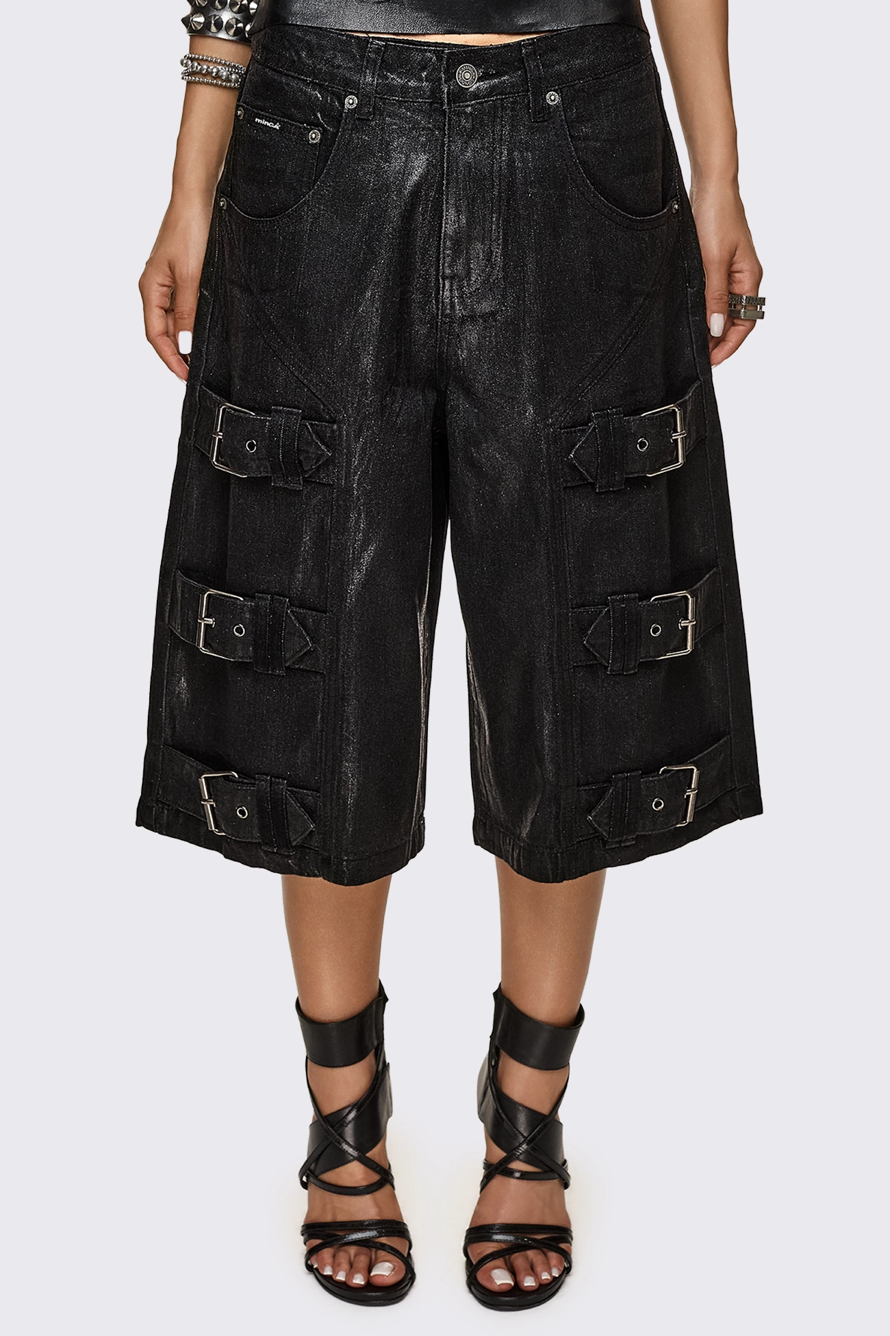 Black coated denim jorts with metallic finish and buckle strap details, longline relaxed fit in a grunge aesthetic.