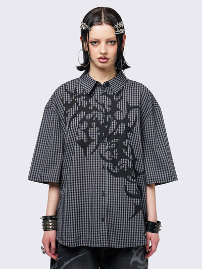 Oversized grunge-style button-up shirt with a dark grey and black checkered pattern and bold black abstract tribal graphic print. The short-sleeve design gives it a relaxed fit, while the edgy pattern adds a rebellious, alternative touch. Styled with studded bracelets for a punk-inspired look.
