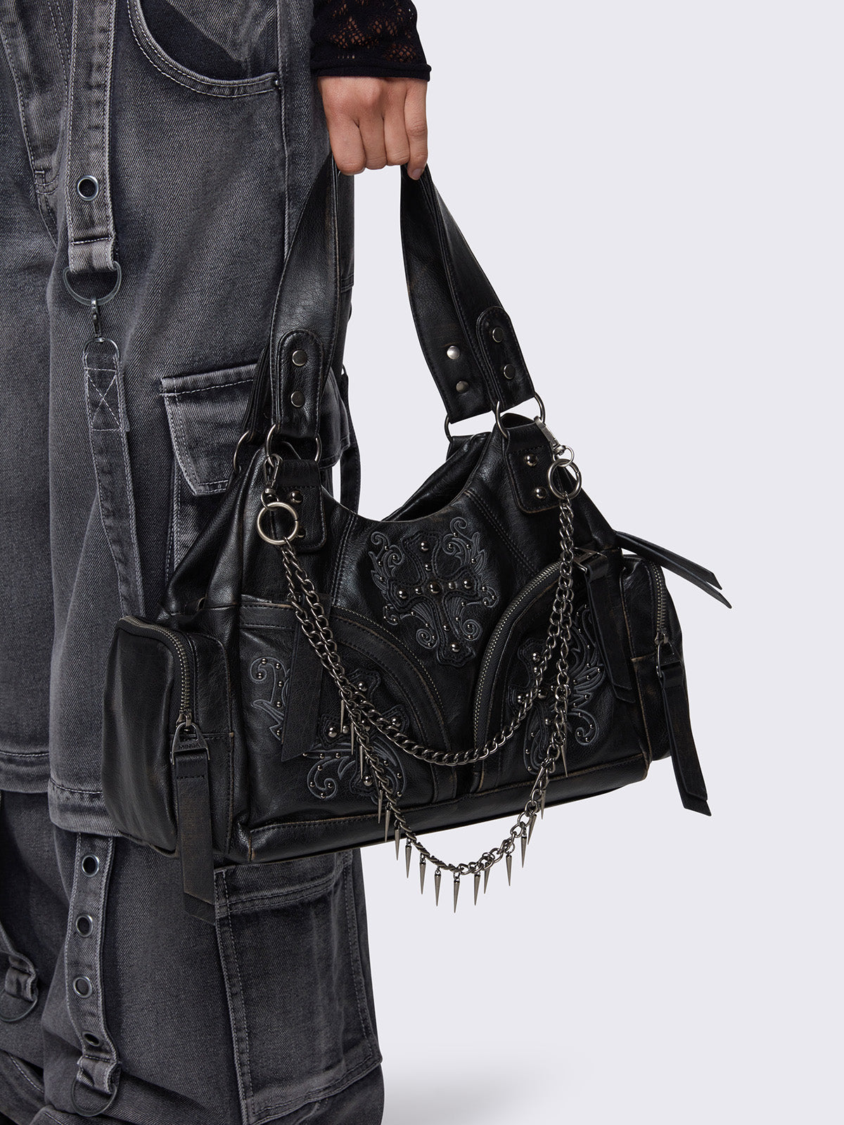 Black faux leather shoulder bag with silver chains, spike charms, side pockets, and embroidered cross detail on the front.