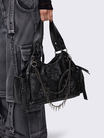 Black faux leather shoulder bag with silver chains, spike charms, side pockets, and embroidered cross detail on the front.