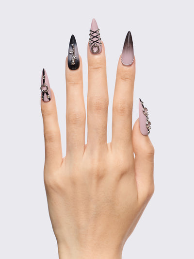 Set of stiletto press-on nails features a bold black and dusty pink ombré base, detailed with metallic piercing accents, faux corset lacing, and 3D charm embellishments. Perfect for adding edge to your outfit without a trip to the salon.