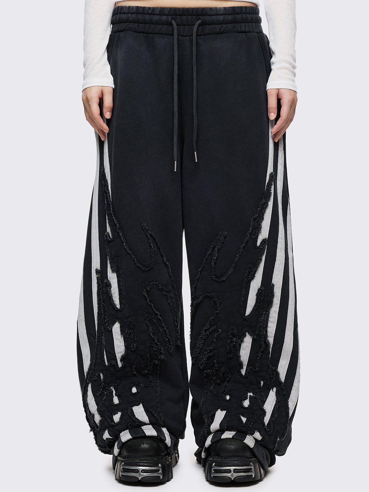 A pair of oversized black wide-leg sweatpants featuring white vertical side stripes and large, black flame-shaped overlays with raw, frayed edges.