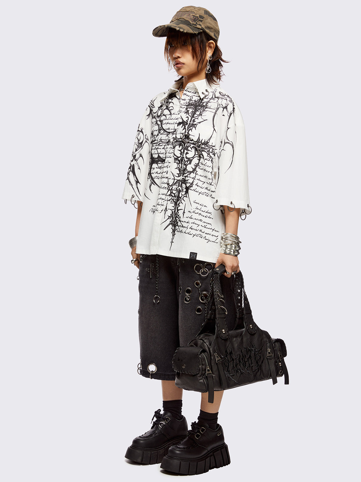 White oversized bowling shirt with black sigilism and lettering graphic print, eyelets with silver O-rings on sleeves and collar and more metal hardware details. 